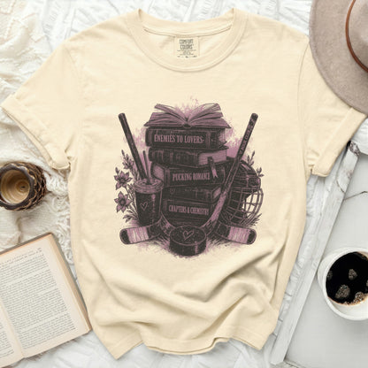Cream t-shirt with books, hockey sticks, and "Enemies to Lovers" text.