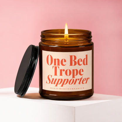 Amber candle with lit wick, lid off, featuring text: One Bed Trope Supporter.
