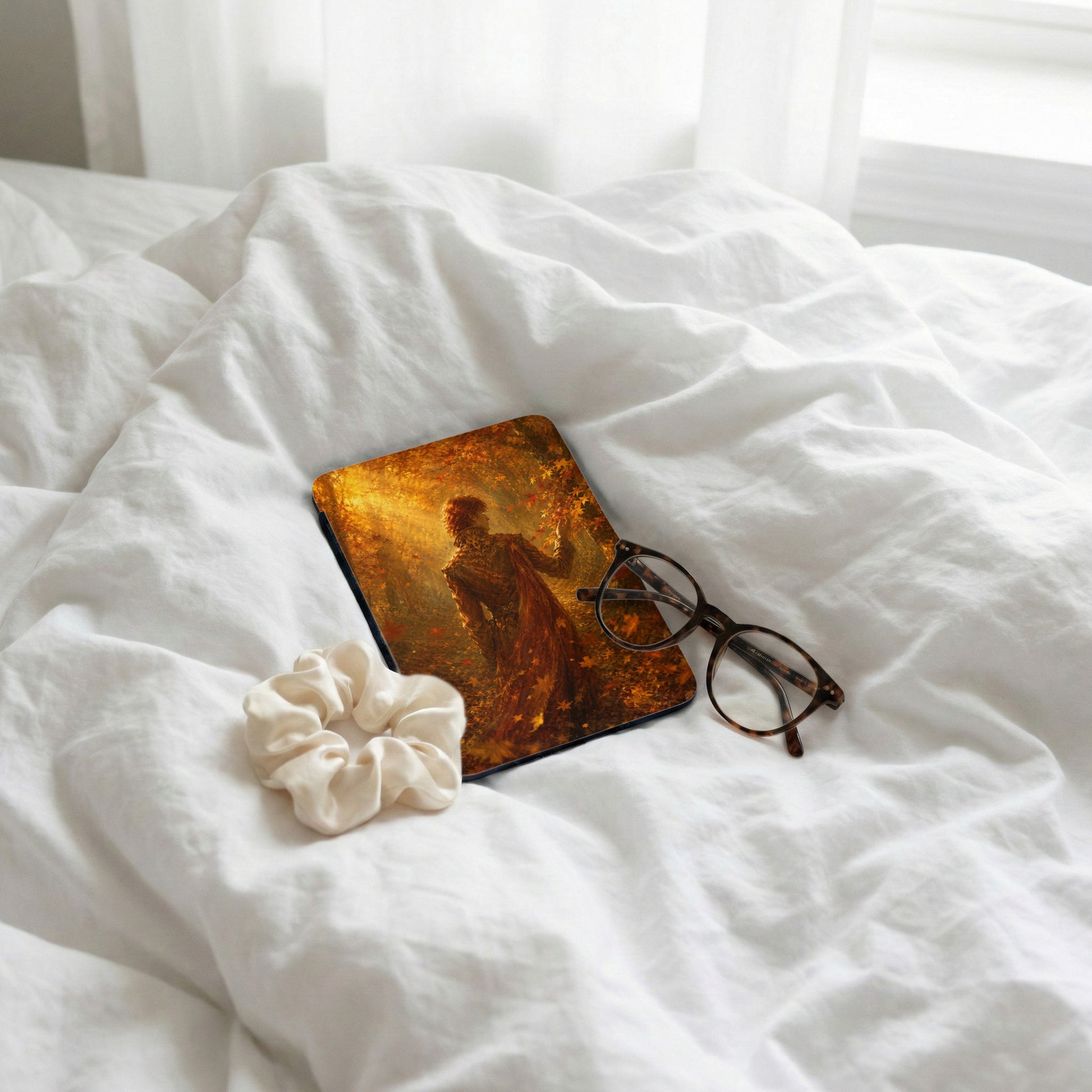 A device with autumnal artwork, glasses, and a scrunchie on white bedding.