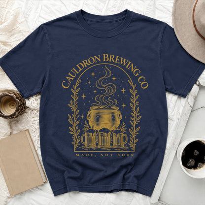 A dark blue t-shirt featuring a gold graphic of a cauldron, beer mugs, stars, and the text CAULDRON BREWING CO.