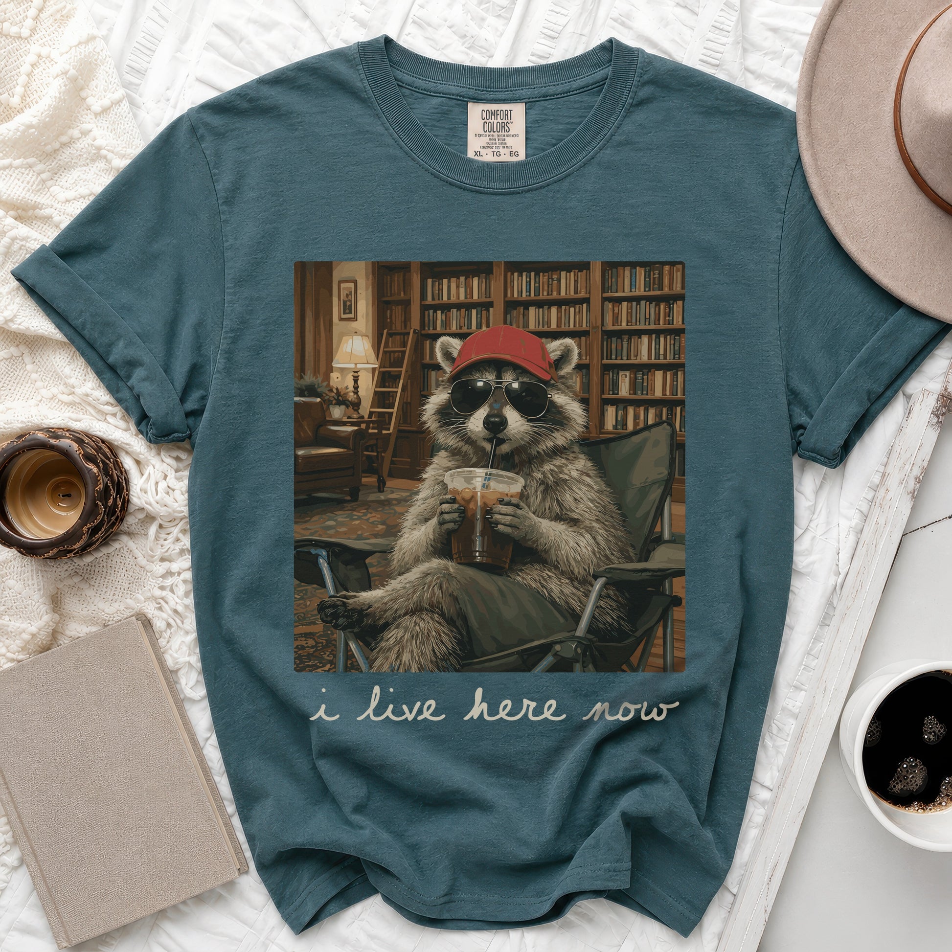 Teal t-shirt with raccoon in sunglasses drinking iced coffee in library graphic.