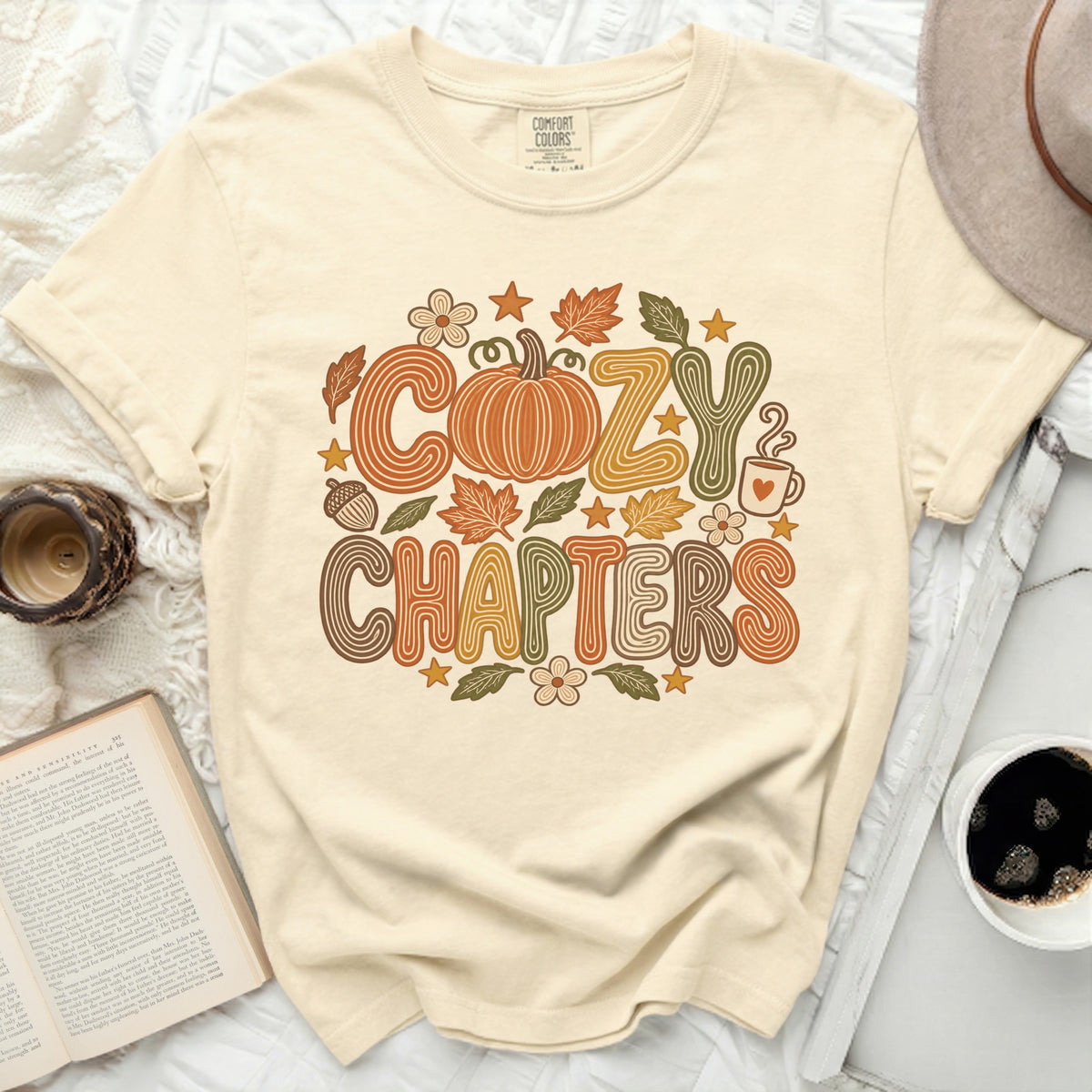 Cream t-shirt with "Cozy Chapters" text, pumpkin, leaves, and coffee cup design.