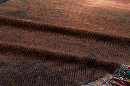 a close-up view of a textured fabric, possibly a rug or textile, with a warm, earthy color palette. The fabric appears to be made of wool or a similar material, with a slightly wrinkled and folded appearance. The image does not contain any discernible text or countable objects, and the focus is on the texture and color of the fabric.