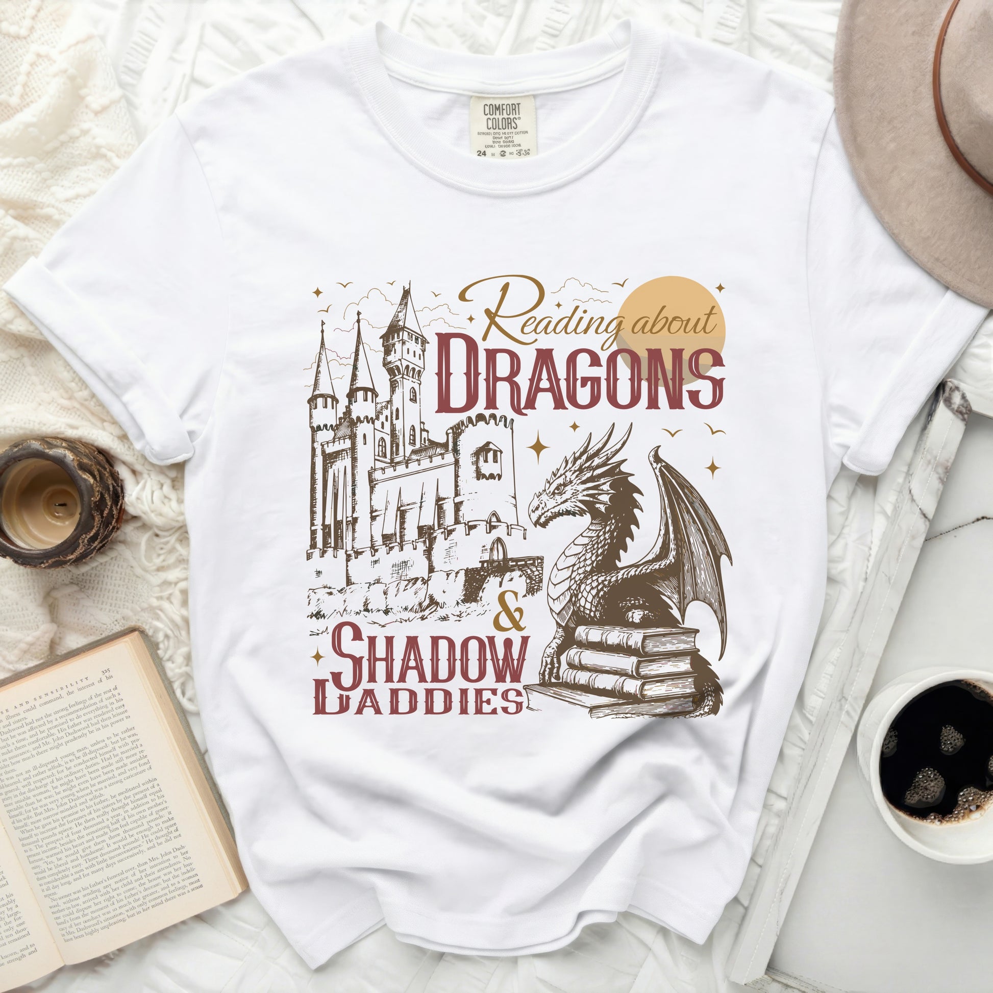 White t-shirt with graphic of a castle, dragon, books, and text "Reading about DRAGONS & SHADOW DADDIES".