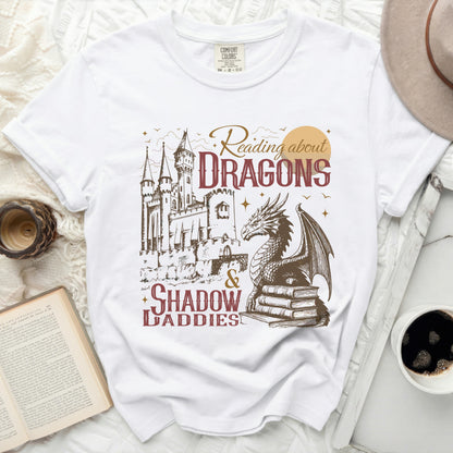 White t-shirt with graphic of a castle, dragon, books, and text "Reading about DRAGONS & SHADOW DADDIES".