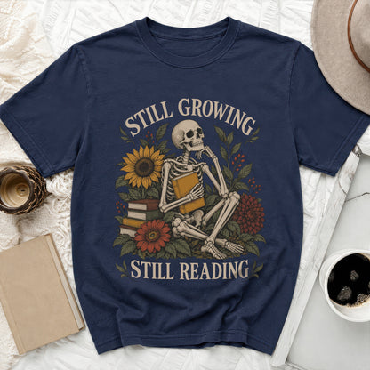 Skeleton sitting with book, surrounded by flowers, with text "STILL GROWING STILL READING".