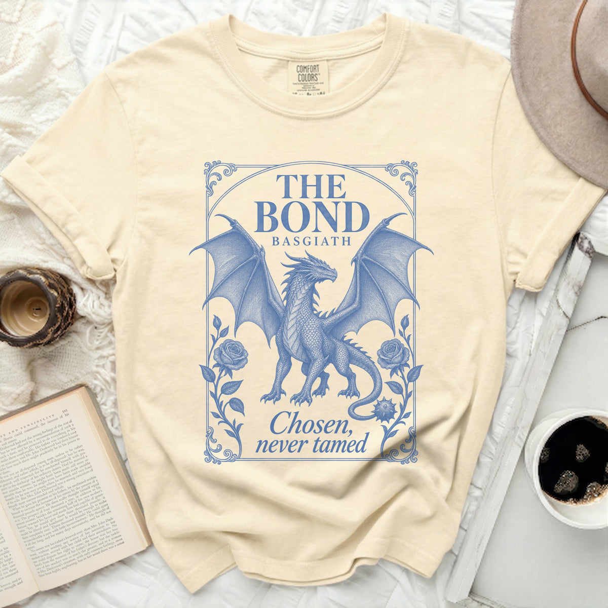 A cream-colored t-shirt featuring a blue graphic of a winged dragon framed by roses and text that reads The Bond Basgiath Chosen, never tamed.