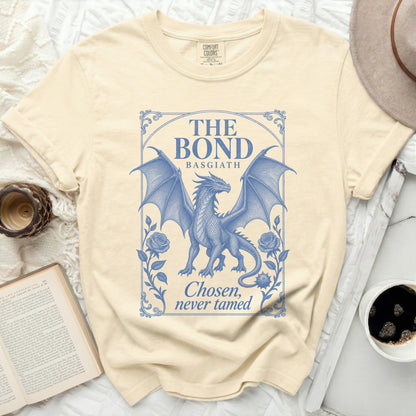 A cream-colored t-shirt featuring a blue graphic of a winged dragon framed by roses and text that reads The Bond Basgiath Chosen, never tamed.
