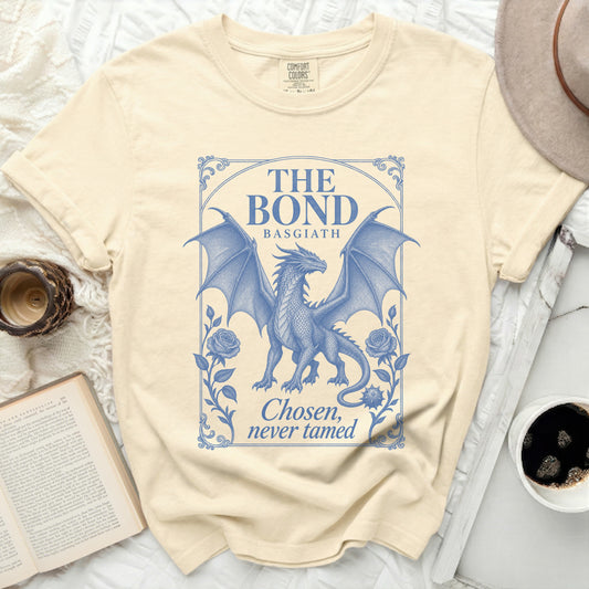 A cream-colored t-shirt featuring a blue graphic of a winged dragon framed by roses and text that reads The Bond Basgiath Chosen, never tamed.
