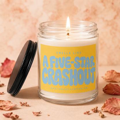 Lit candle with yellow label reading "Smells Like A Five-Star Crashout".