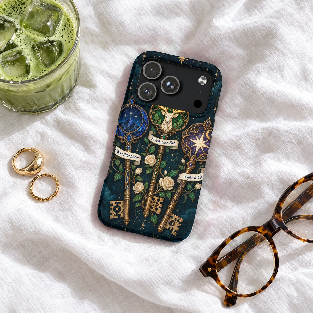 A dark blue phone case with three ornate keys featuring quotes, resting on a white blanket with glasses, rings, and iced matcha.