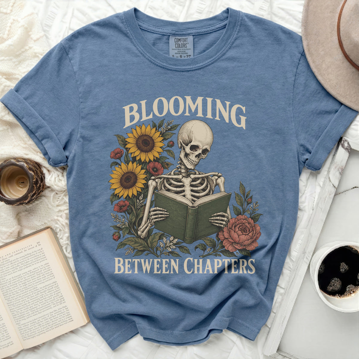 Blue t-shirt with skeleton reading book surrounded by flowers, text "BLOOMING BETWEEN CHAPTERS".
