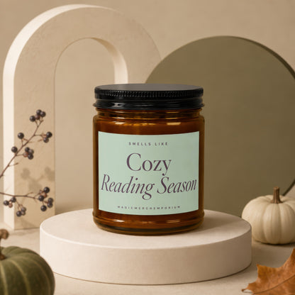 Amber jar candle with "Cozy Reading Season" label on a pedestal, featuring decorative pumpkins.