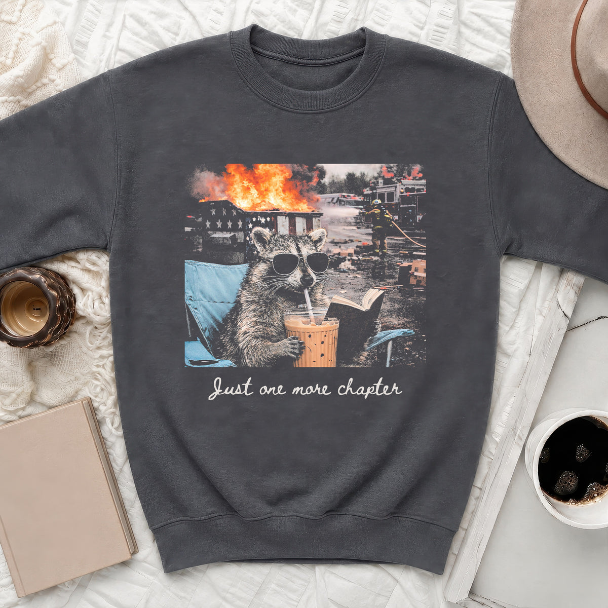 Raccoon in sunglasses drinks from a cup while reading a book with a fire in the background.