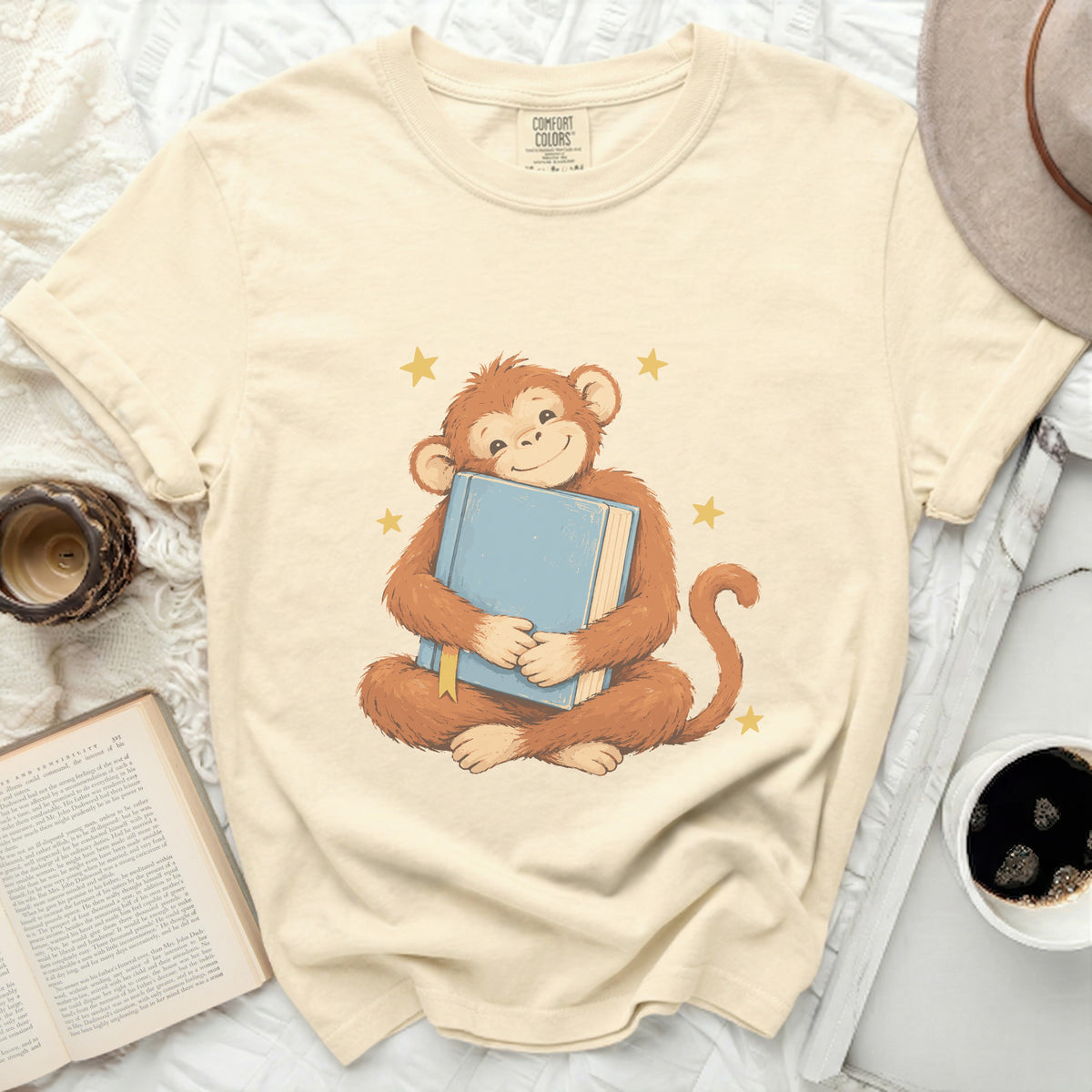 Cream t-shirt with a cheerful monkey hugging a blue book surrounded by stars.