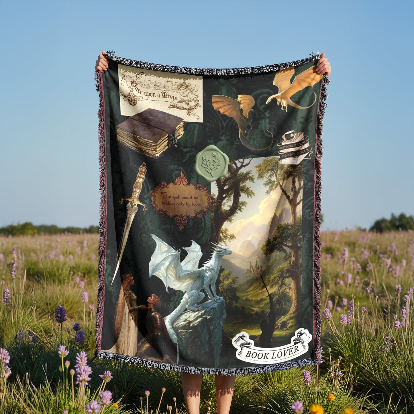 A person is holding a large blanket with a dragon and various fantasy-themed illustrations, including a book, a sword, and a castle, in a field with purple flowers.