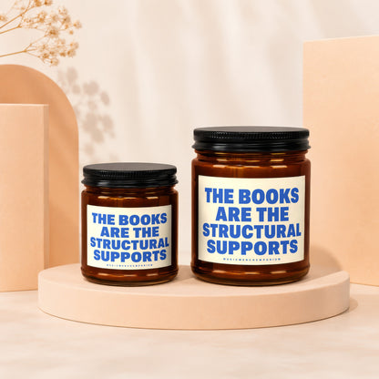 Two amber glass candles with black lids, featuring labels that read "The Books Are The Structural Supports."