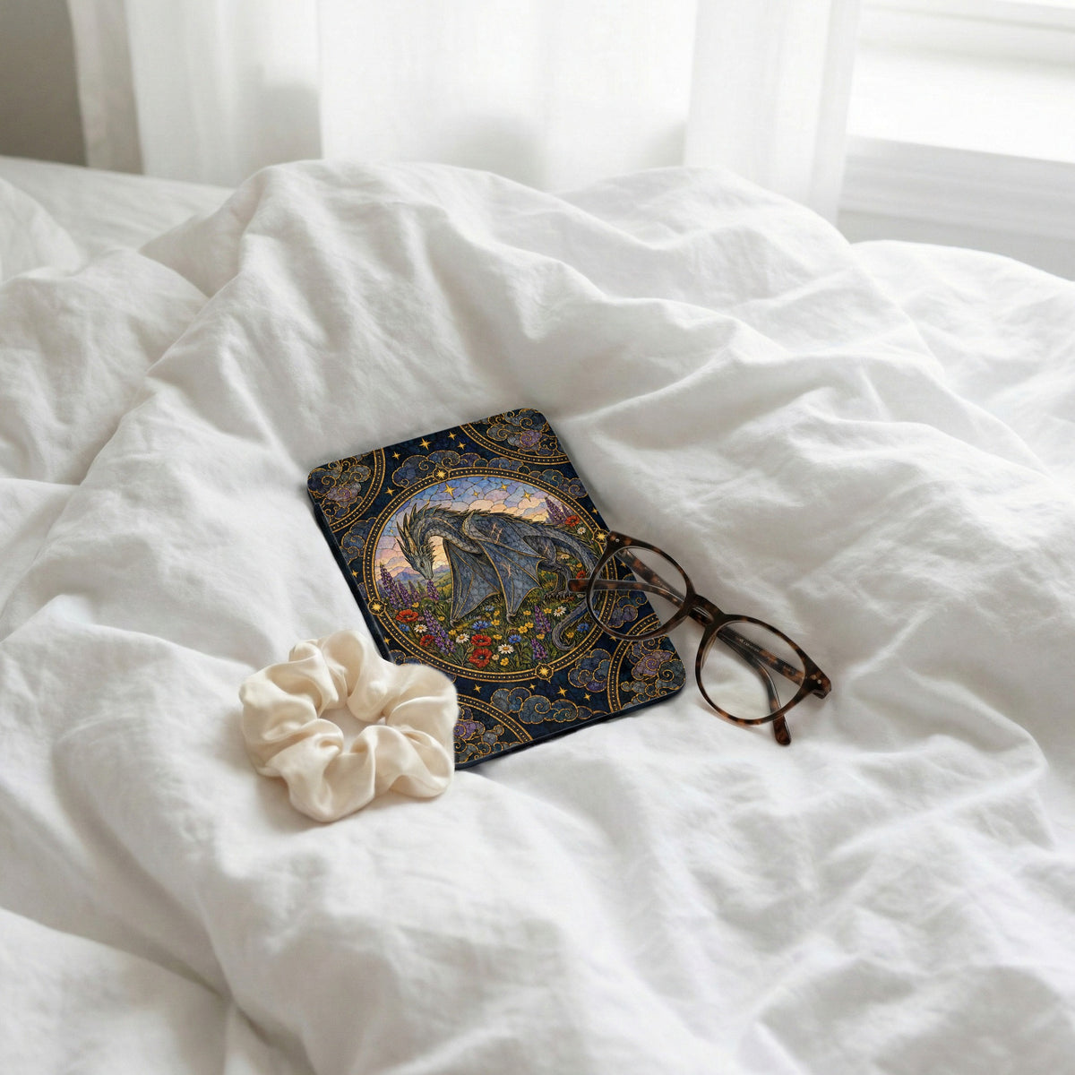 A patterned Kindle case featuring a dragon rests on a white bed with glasses and a scrunchie.