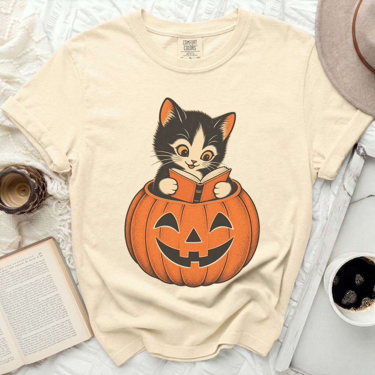 Black kitten reading a book inside a jack-o'-lantern graphic on a cream t-shirt.