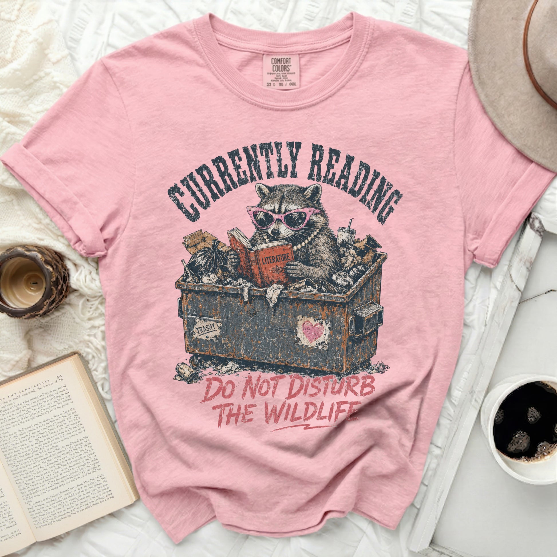 Pink t-shirt with raccoon in trash can reading a book.