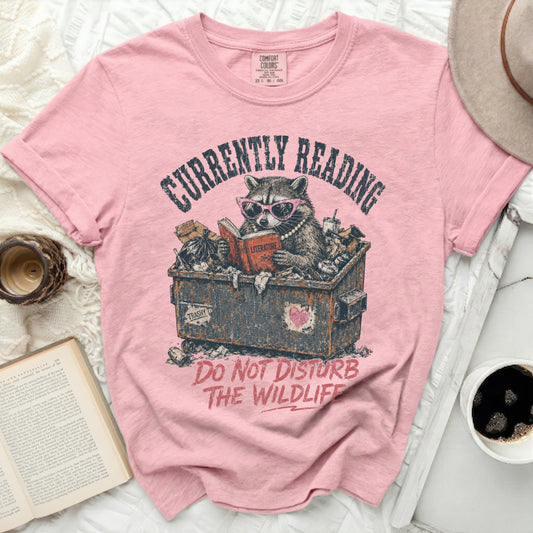Pink t-shirt with raccoon in trash can reading a book.