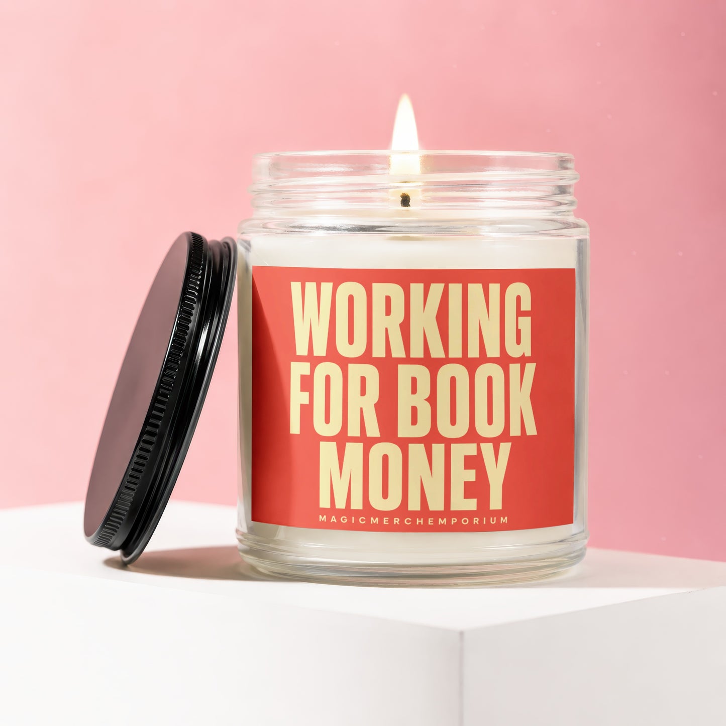 Glass jar candle with lit wick and black lid, saying "Working for Book Money".
