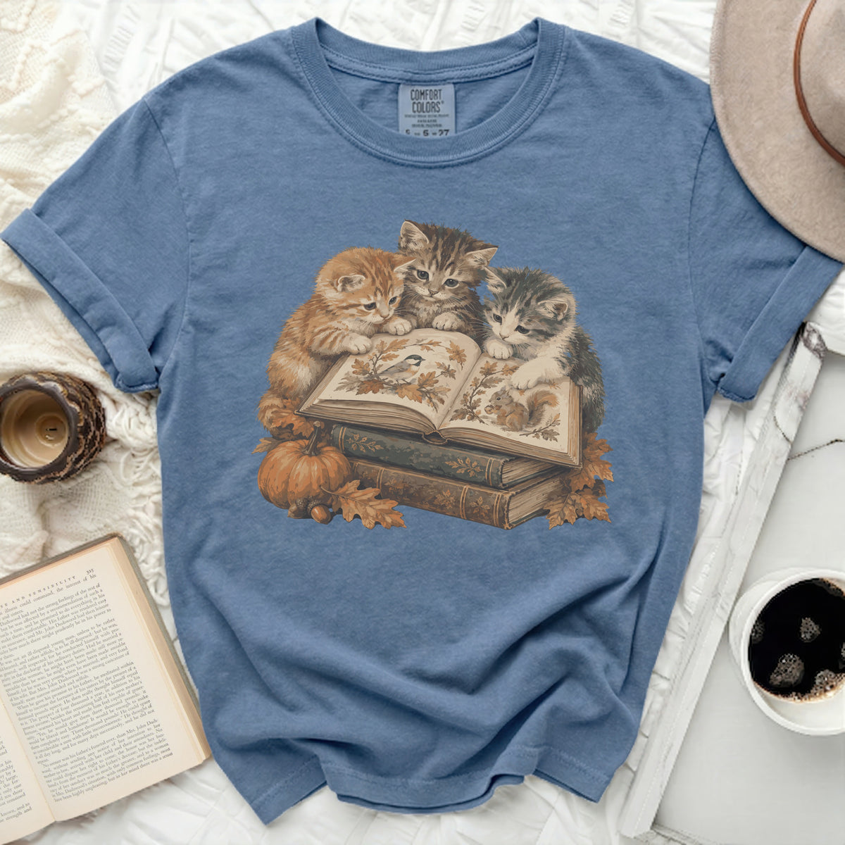 Three kittens read a book surrounded by autumn leaves and pumpkins.