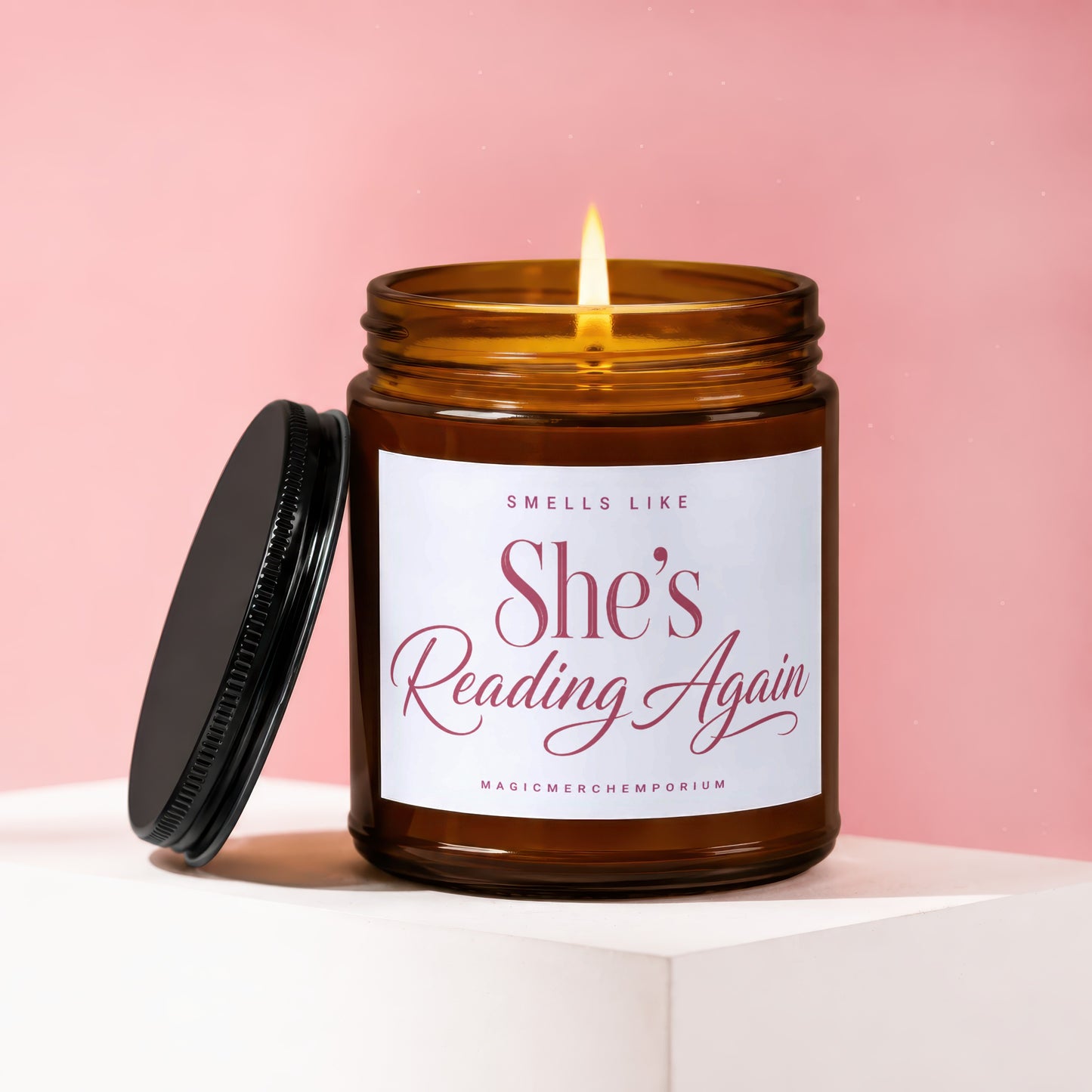 Amber glass candle with lit flame, black lid off, and label reading "She's Reading Again".