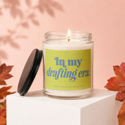 Glass candle burning with a lid off, featuring text "In my drafting era."