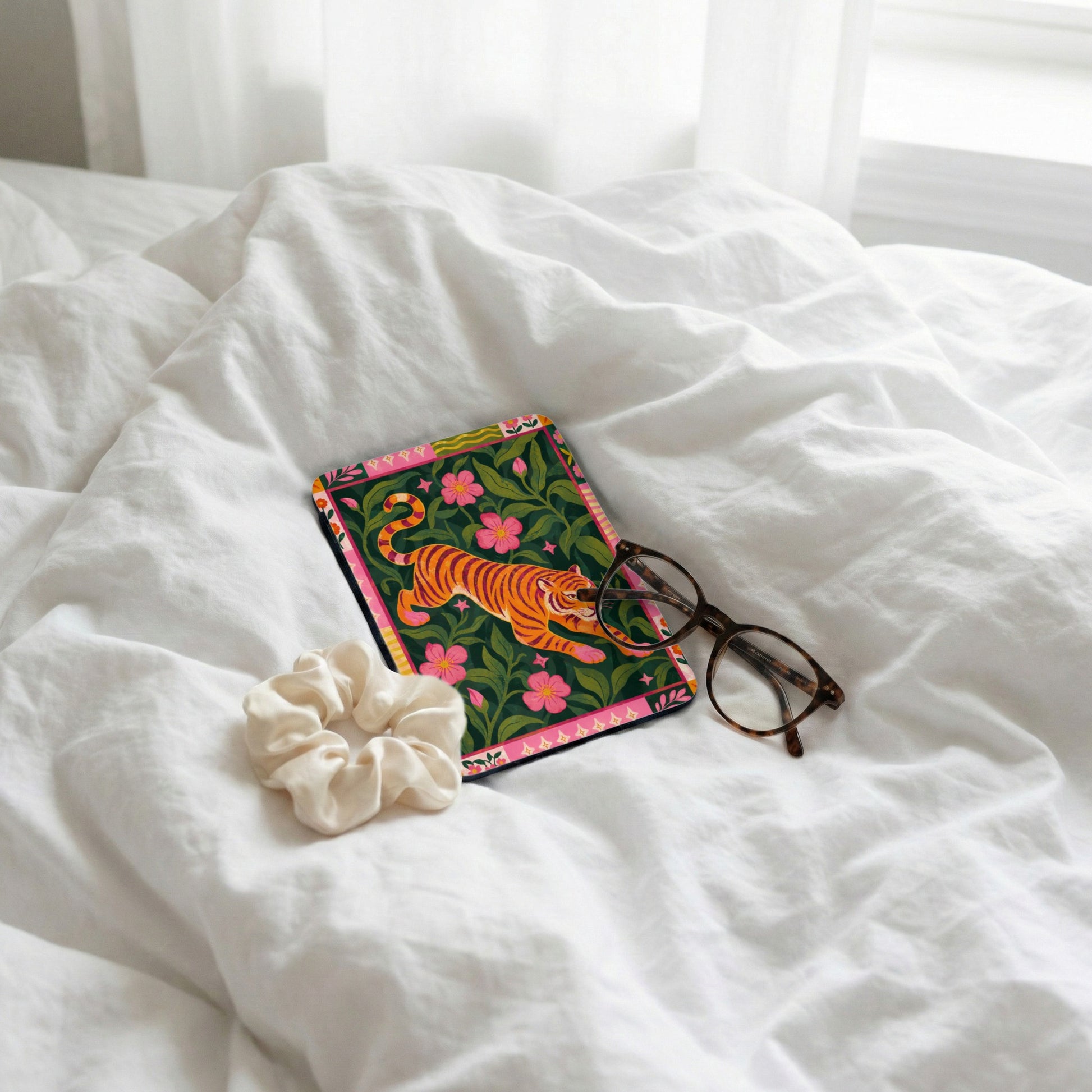 Card with tiger and floral design next to glasses and scrunchie on white bedding.