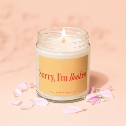 Glass jar candle with lit wick and text "Sorry, I'm Booked" surrounded by petals.