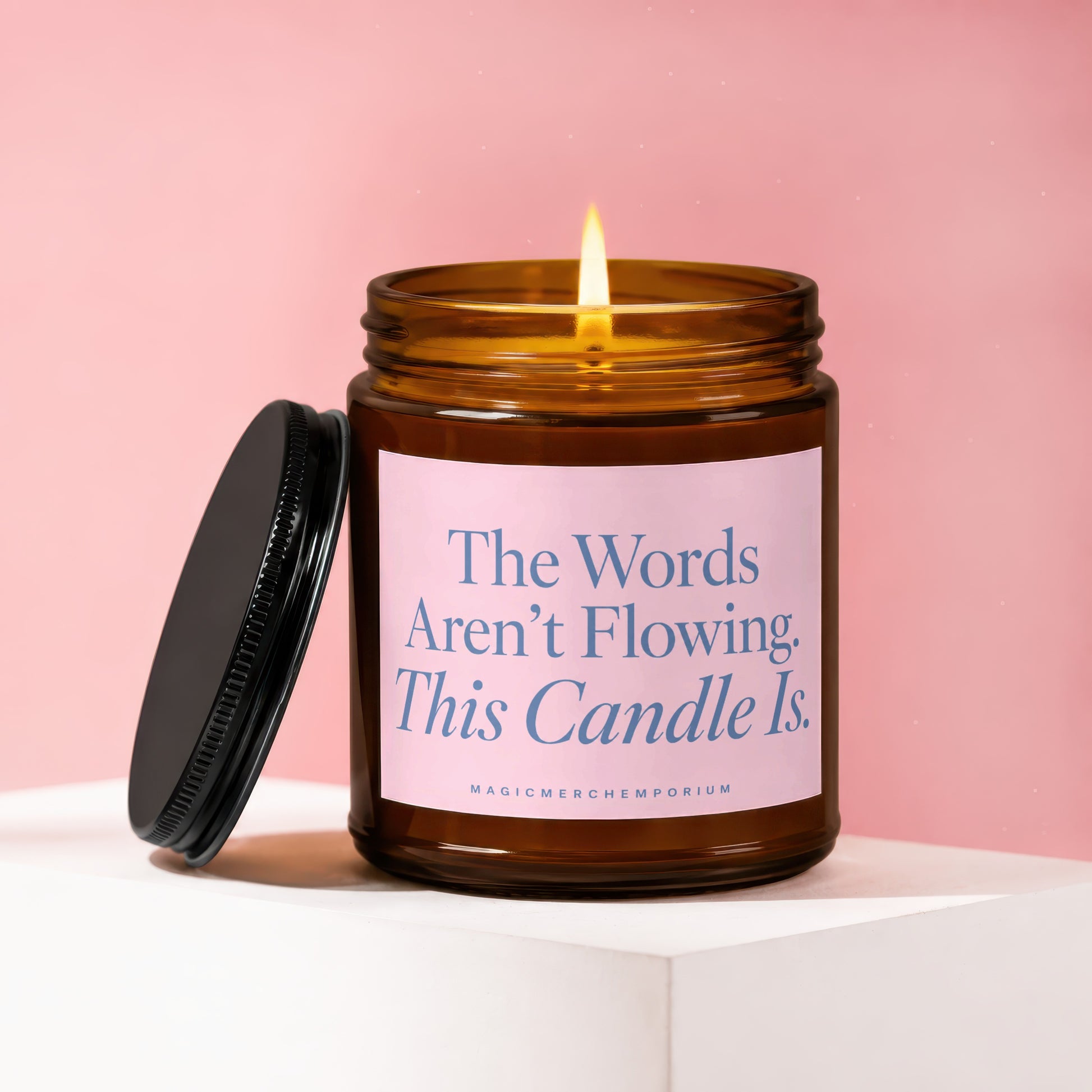 Amber glass candle with a burning wick and pink label reading "The Words Aren't Flowing. This Candle Is."