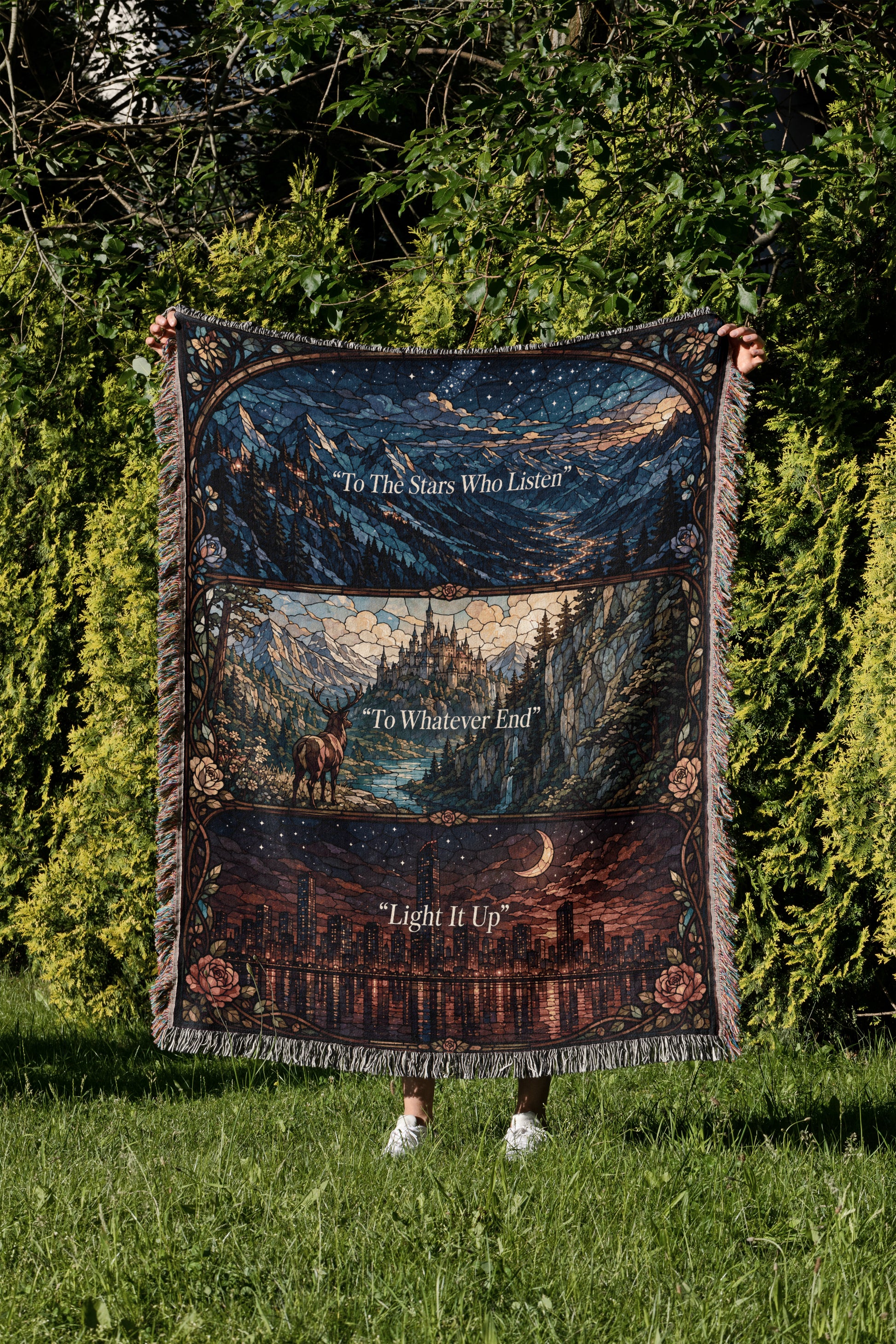 A person is holding a large, colorful blanket with a mountain landscape and a city skyline printed on it, standing in a grassy area with trees in the background.