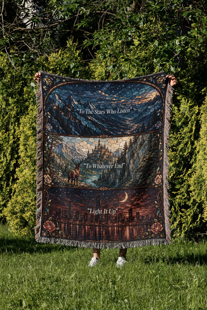 A person is holding a large, colorful blanket with a mountain landscape and a city skyline printed on it, standing in a grassy area with trees in the background.