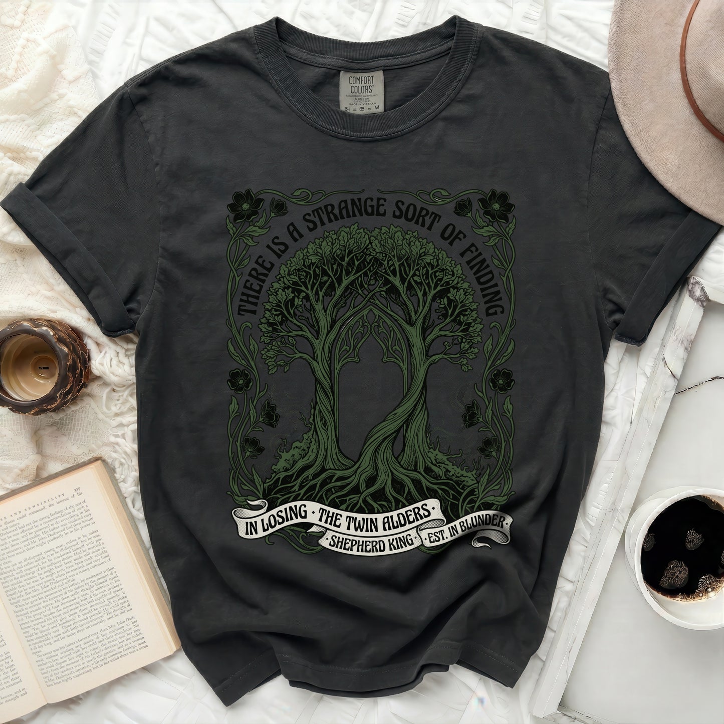 Dark t-shirt with green trees and text "There is a strange sort of finding".