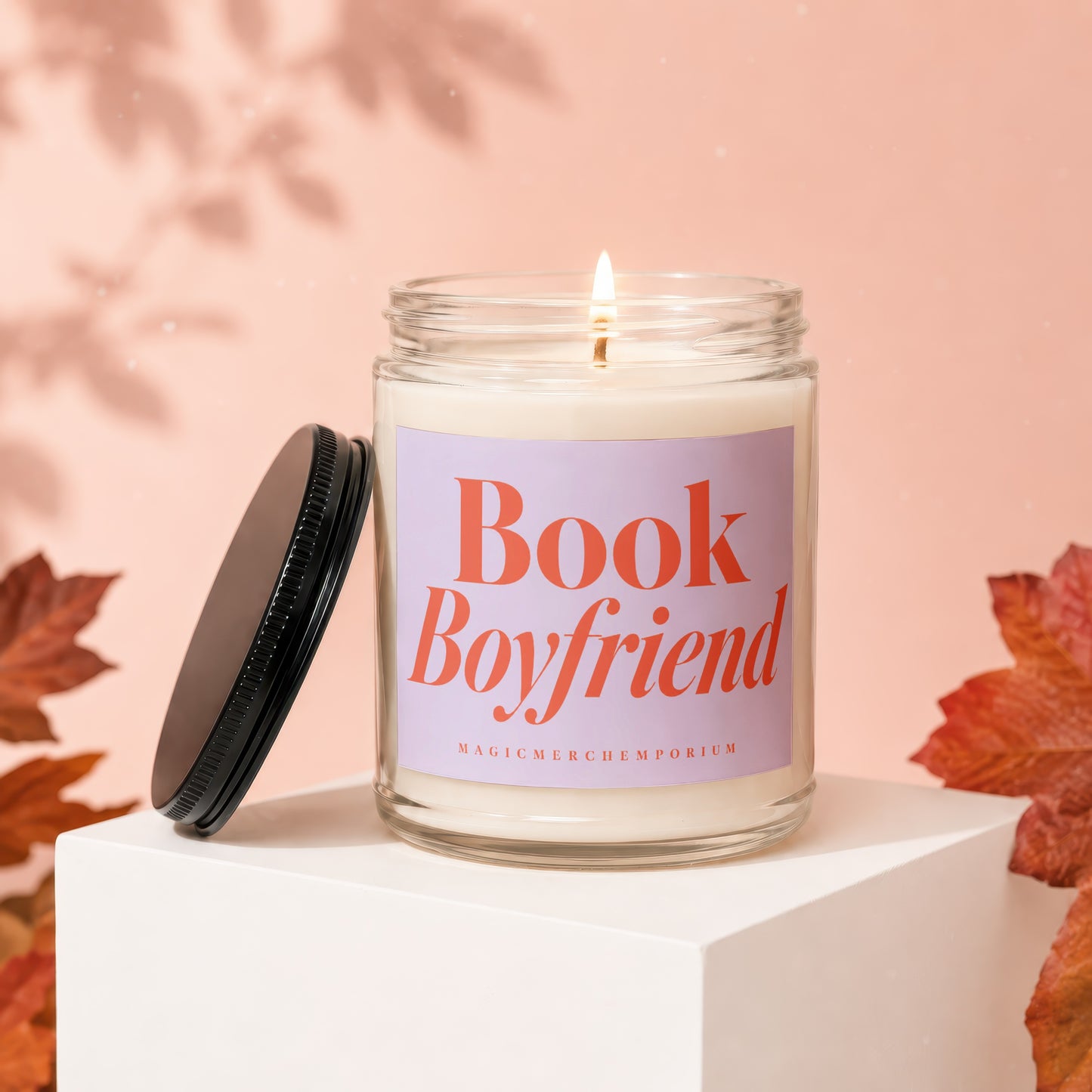 Candle with "Book Boyfriend" text, lid to the side, on a white pedestal with autumn leaves.