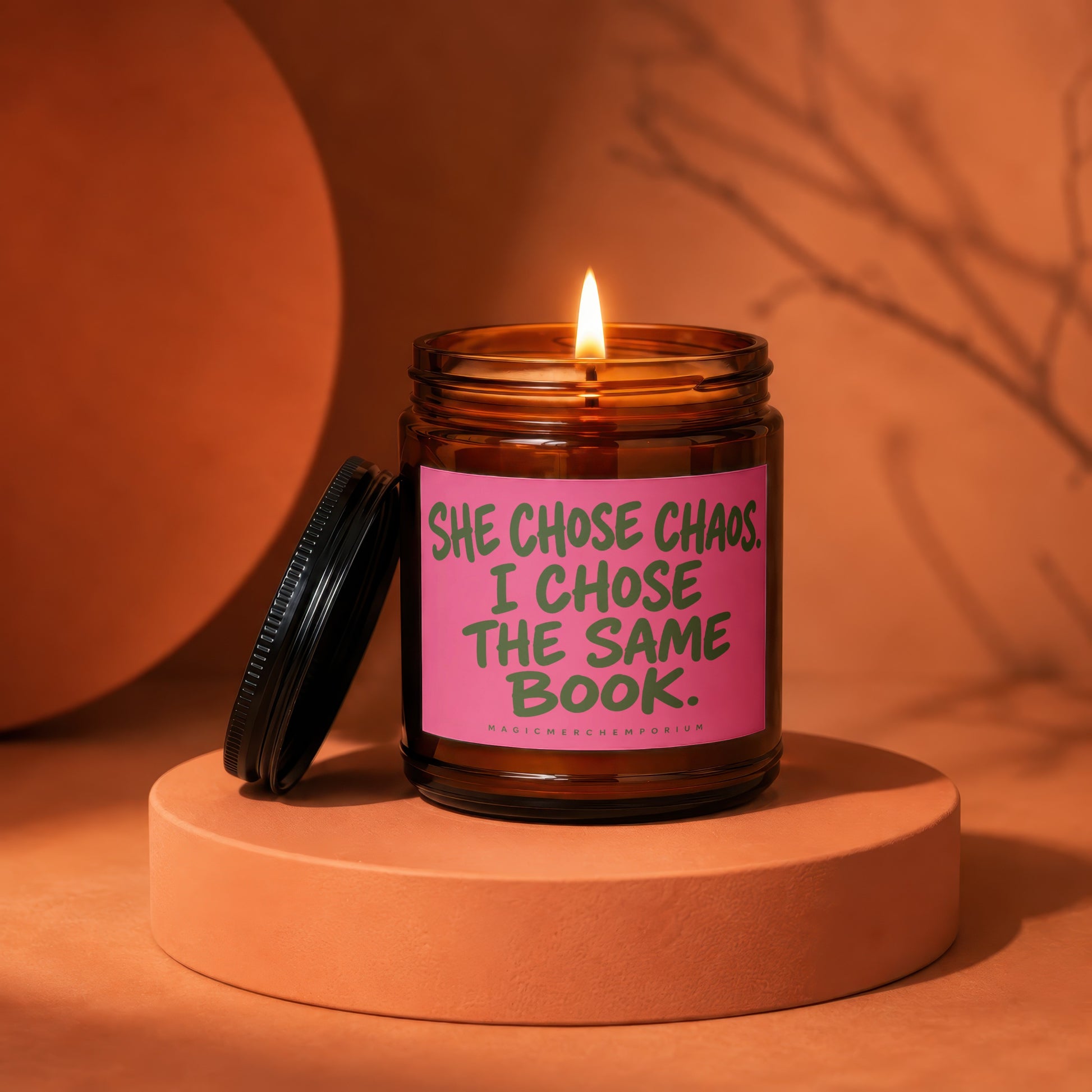 Brown candle in amber jar with pink label reading "SHE CHOSE CHAOS. I CHOSE THE SAME BOOK."