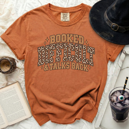 Orange t-shirt with text "Booked, Difficult & Talks Back" in distressed font.