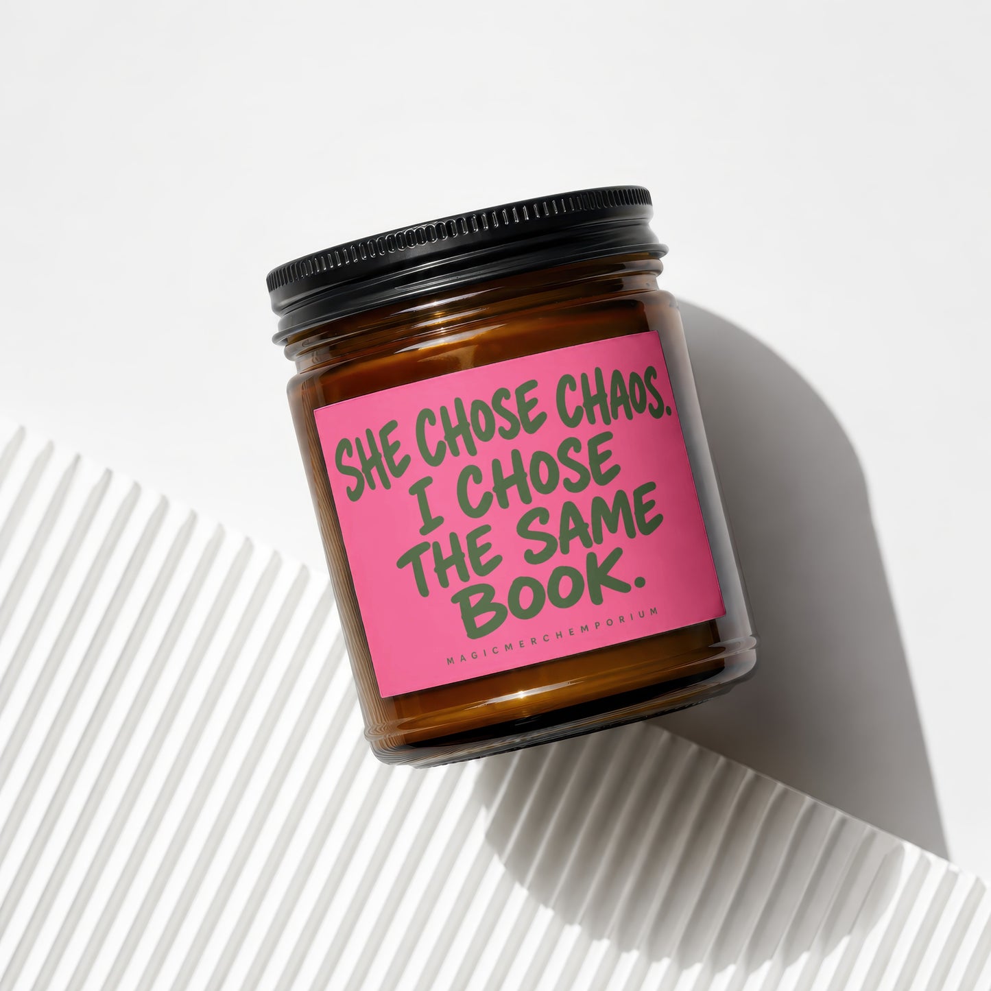 Brown glass candle with pink label reading "SHE CHOSE CHAOS. I CHOSE THE SAME BOOK."