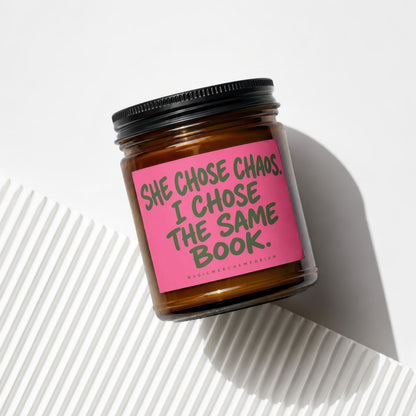 Brown glass candle with pink label reading "SHE CHOSE CHAOS. I CHOSE THE SAME BOOK."
