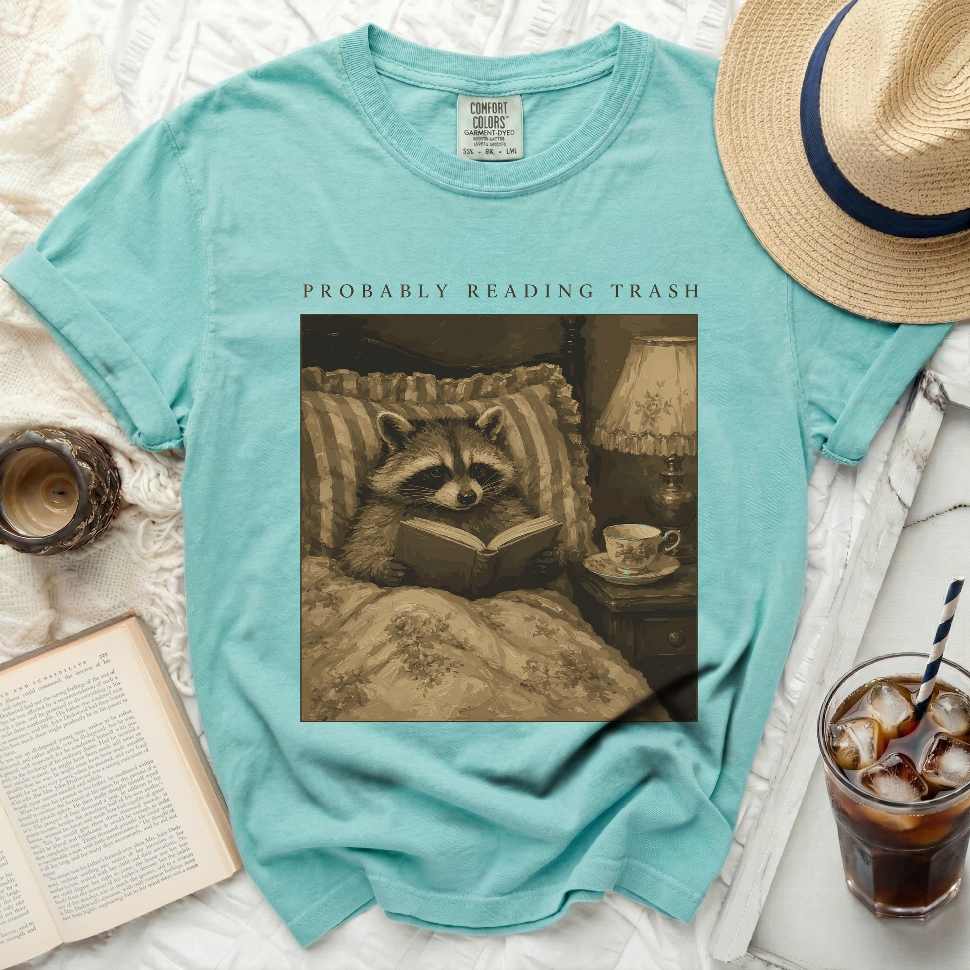 Teal t-shirt with graphic of a raccoon reading in bed, "PROBABLY READING TRASH" text.