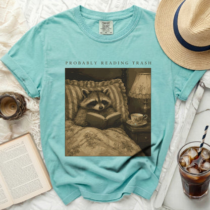 Teal t-shirt with graphic of a raccoon reading in bed, "PROBABLY READING TRASH" text.