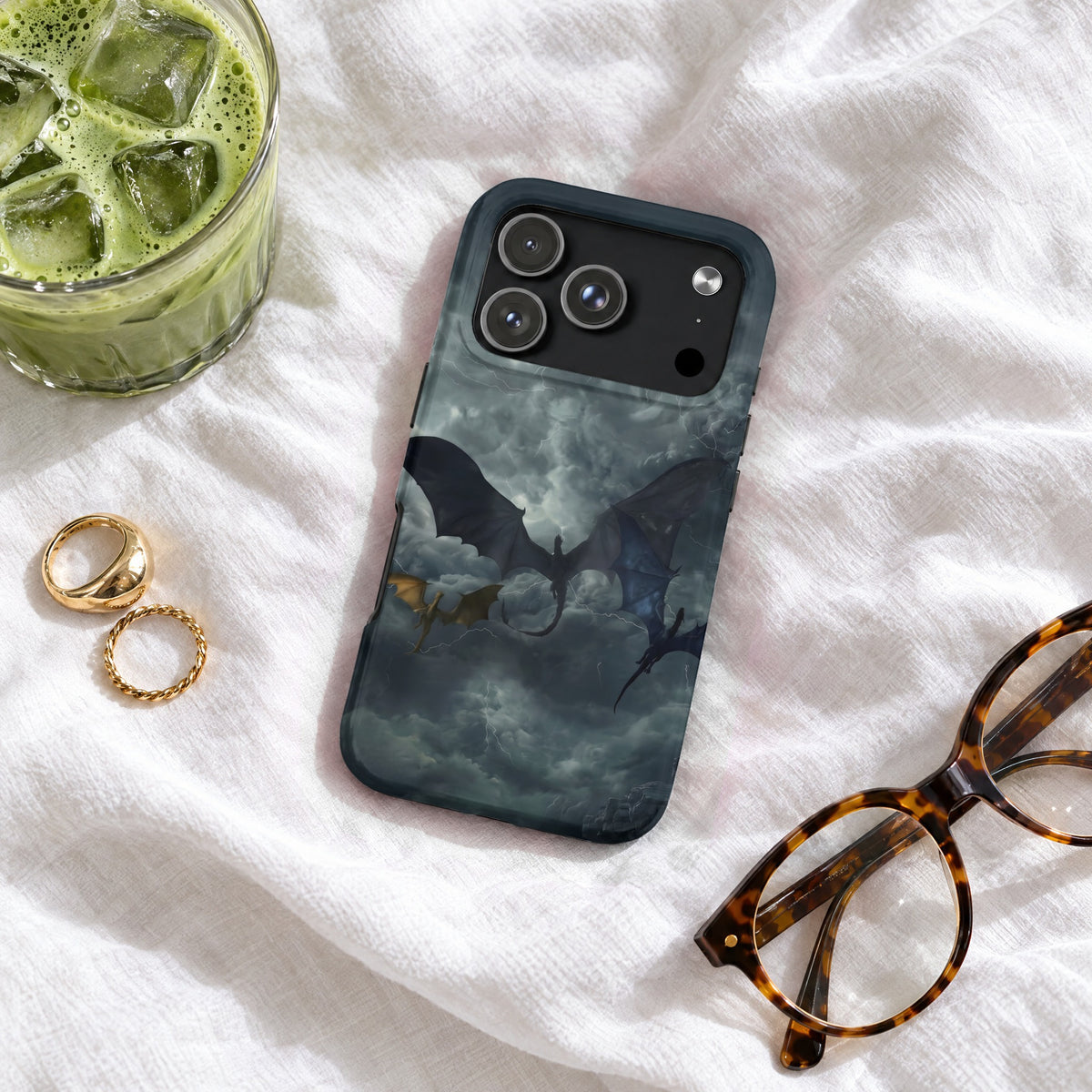 A smartphone case featuring a dragon graphic resting on white fabric near glasses and a drink.