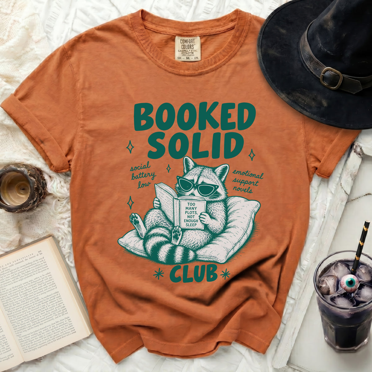 Orange t-shirt with a raccoon reading a book and text "BOOKED SOLID CLUB".