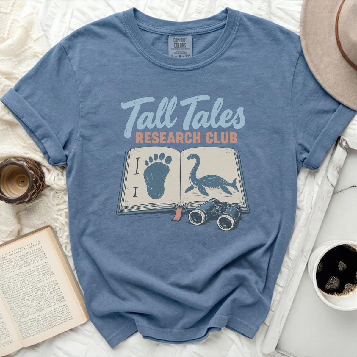 Blue t-shirt with "Tall Tales Research Club" and illustrations of a book, footprint, and Loch Ness Monster.