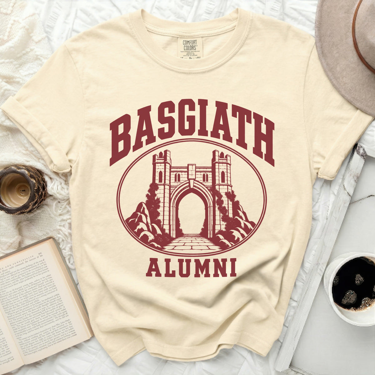 A cream crewneck t-shirt featuring a maroon collegiate-style design with the text "Basgiath Alumni" and a stone gateway.