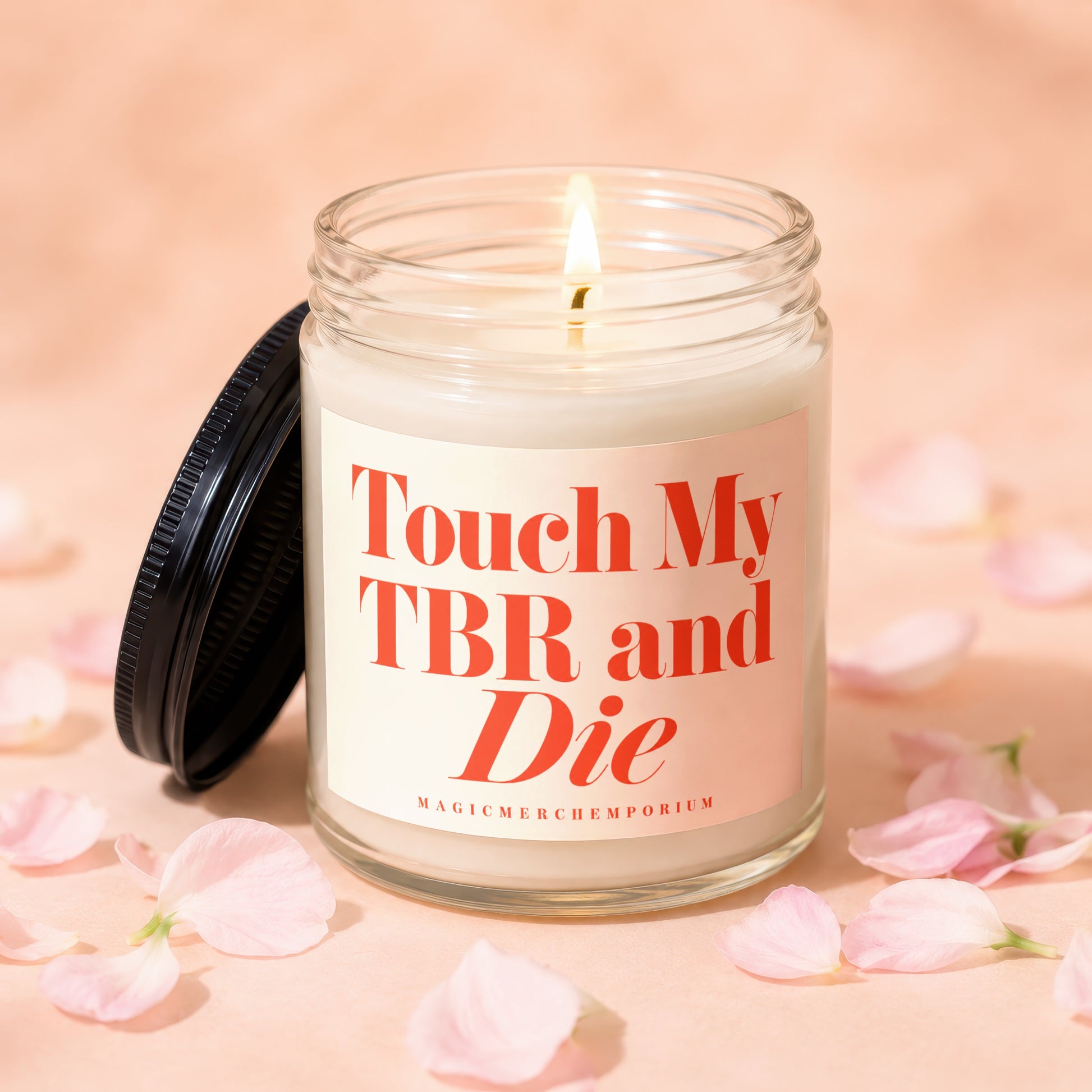 Glass candle with lit wick and black lid displays "Touch My TBR and Die," surrounded by pink petals.