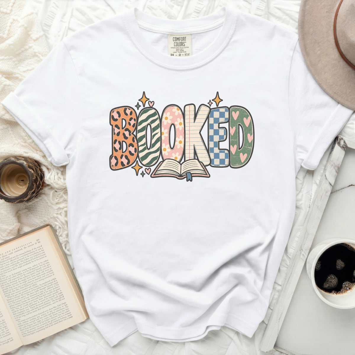 White t-shirt with "BOOKED" graphic in colorful patterned letters over an open book.