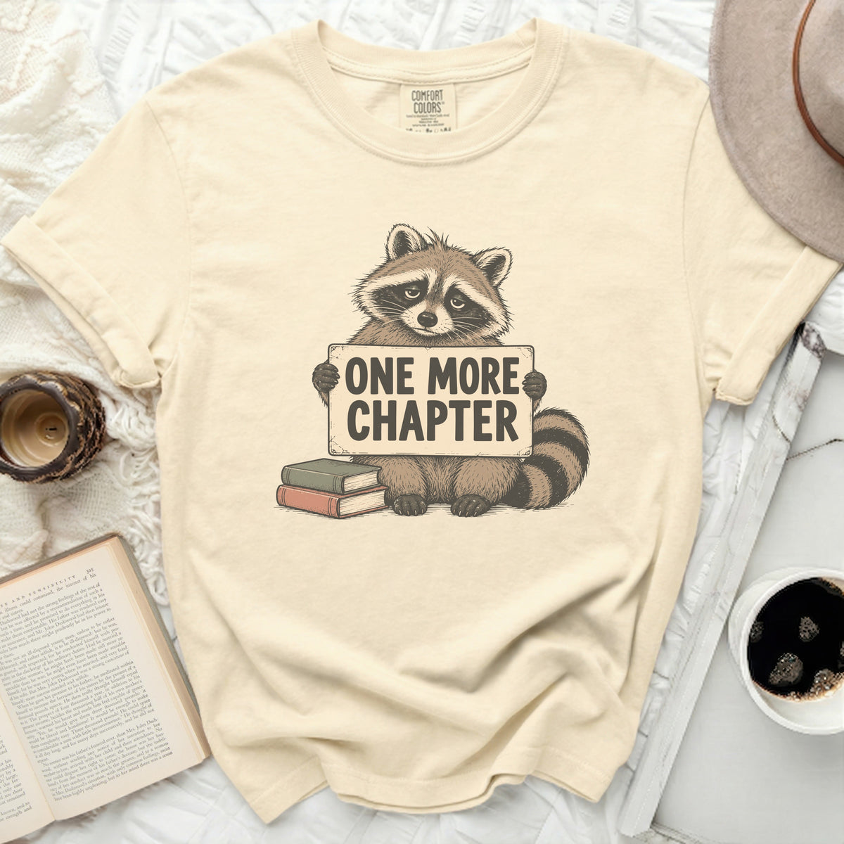 T-shirt with a raccoon holding a sign that says "ONE MORE CHAPTER".