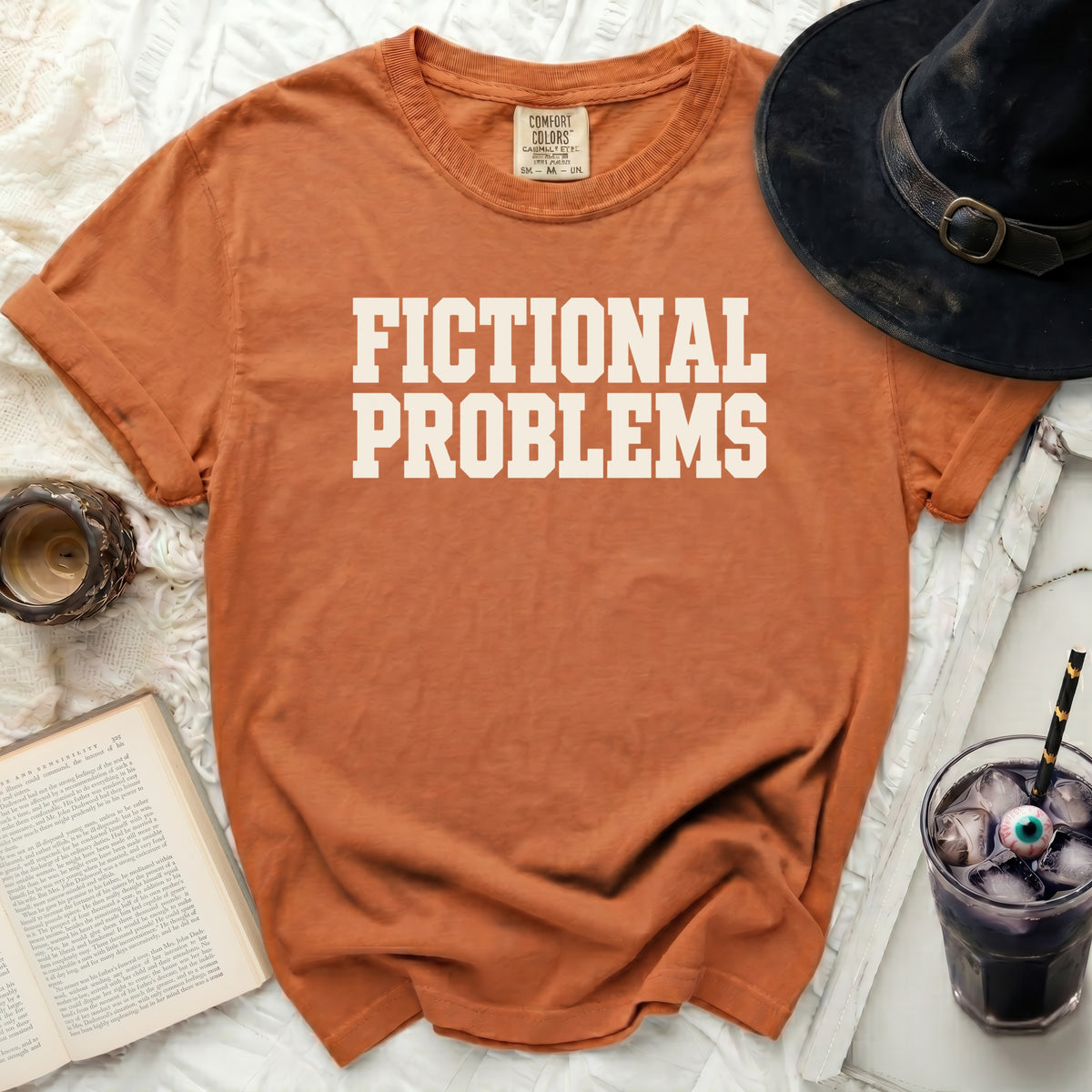 Orange t-shirt with "FICTIONAL PROBLEMS" text, next to a black hat and drink.